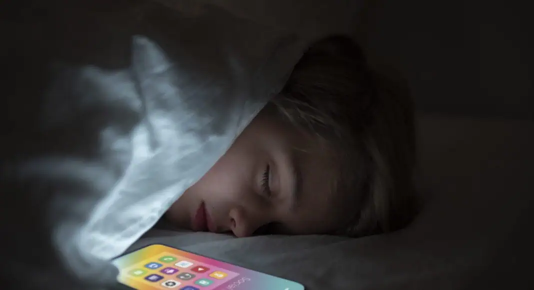 Un bambino dorme sotto una coperta accanto a uno smartphone luminoso con l'app TikTok aperta, la cui luce illumina la stanza buia.
