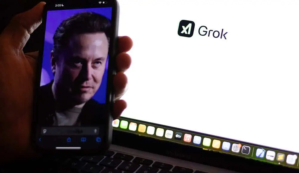 Un telefono mostra il volto di un uomo davanti allo schermo di un computer portatile che visualizza il logo Grok e il testo Grok.