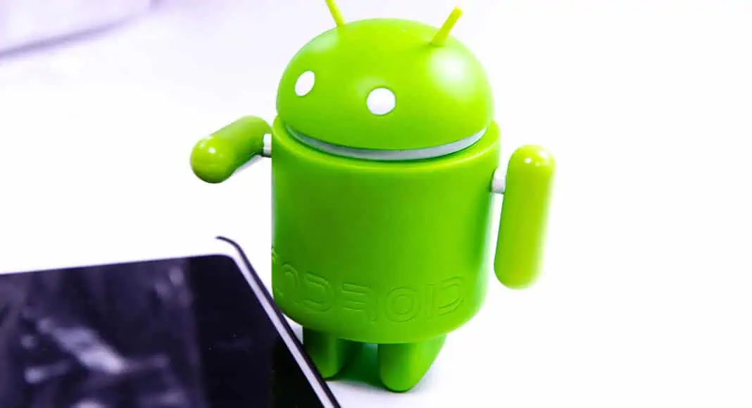 La figura della mascotte verde di Android, nota con il nome in codice Android 17, si trova vicino all'angolo di uno smartphone nero su sfondo bianco.