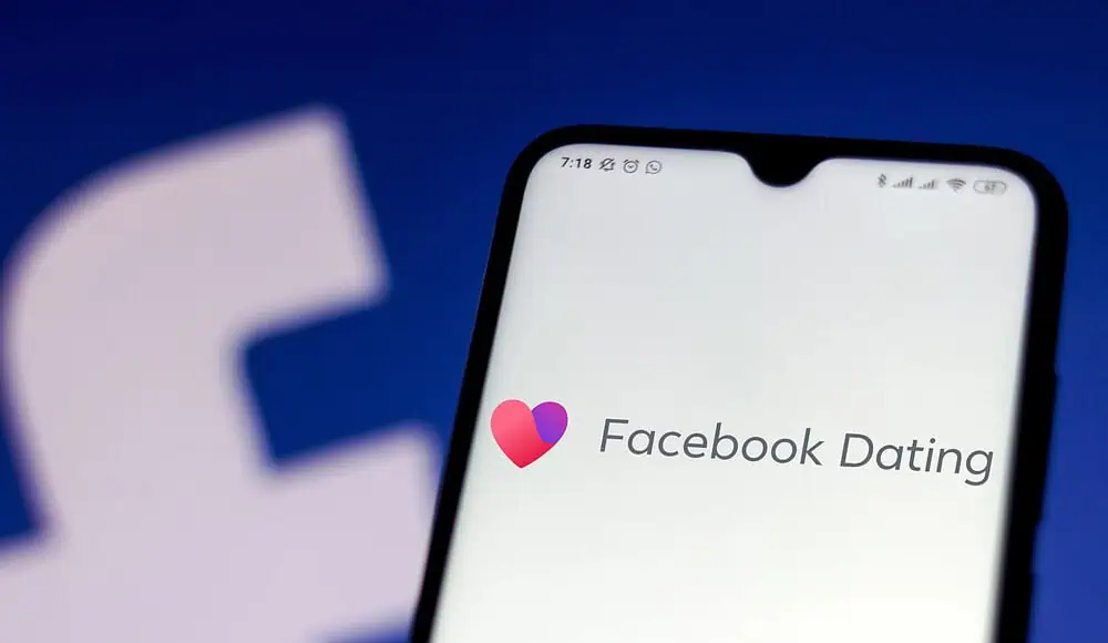 Uno smartphone mostra il logo di Facebook Dating, con un logo di Facebook sfocato sullo sfondo, evidenziando il legame tra Facebook e la sua piattaforma di incontri.