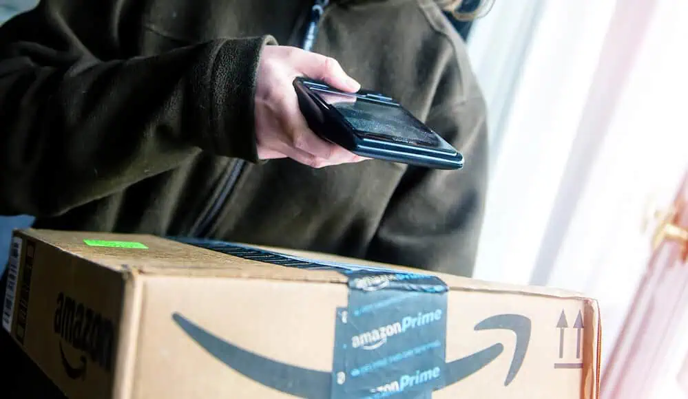 Una persona tiene in mano un pacco Amazon Prime e lo scansiona con uno smartphone vicino a una finestra, pronta a godersi la sua ultima consegna Amazon.