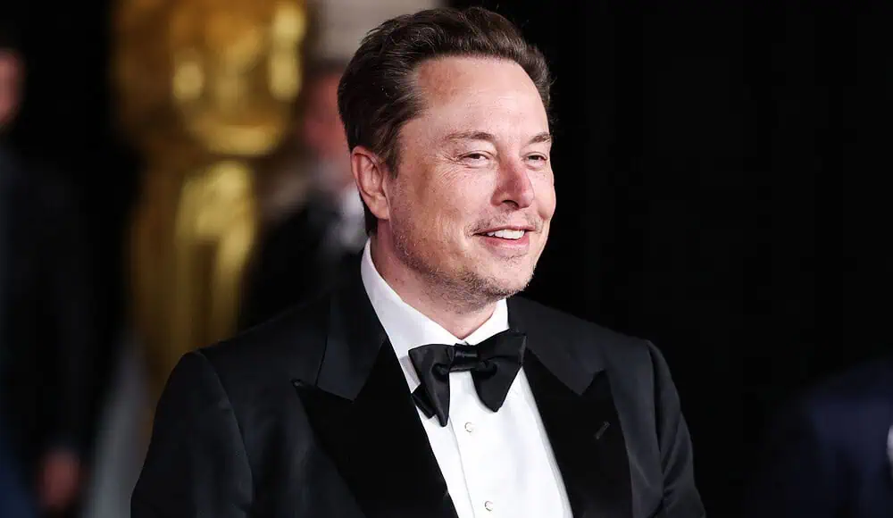 Elon Musk in smoking nero e papillon sorride a un evento formale, con una statua d'oro sullo sfondo.