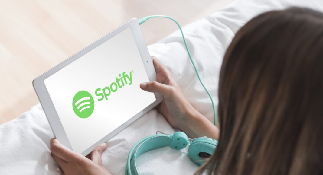 Donna che tiene in mano un tablet con l'app Spotify aperta, con accanto delle cuffie color verde acqua su un letto bianco.