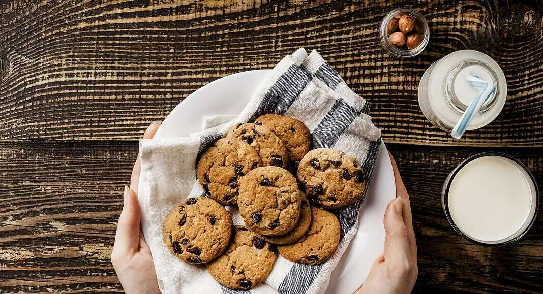 Un piatto di biscotti con gocce di cioccolato fatti in casa è appoggiato su un asciugamano, accanto a bicchieri di latte e a una ciotolina di nocciole, per uno spuntino perfetto a base di biscotti.