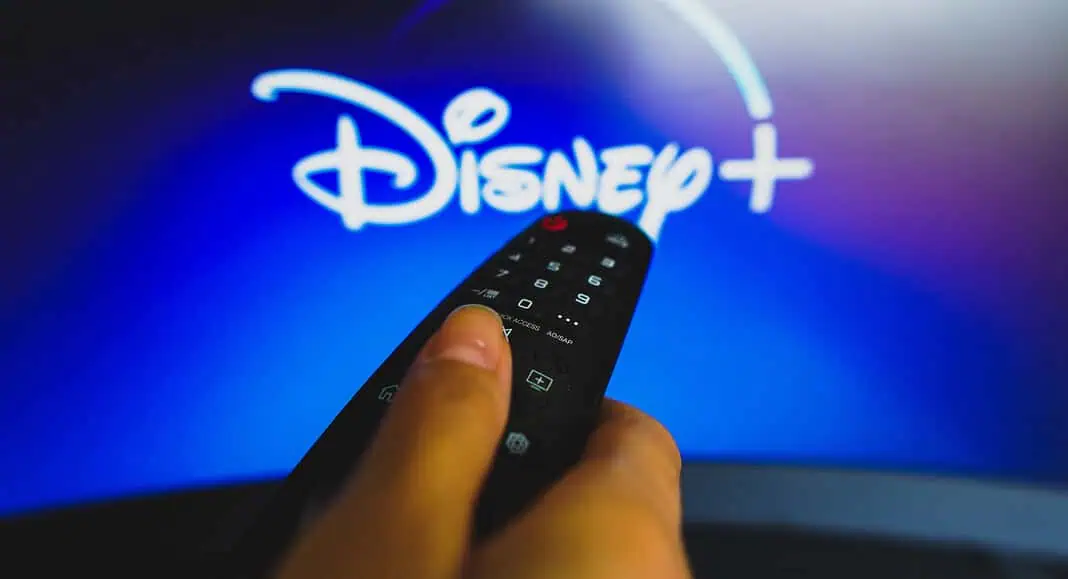 Una mano tiene un telecomando davanti a uno schermo televisivo con il logo Disney+, pronta a trasmettere i programmi preferiti su Disney+.