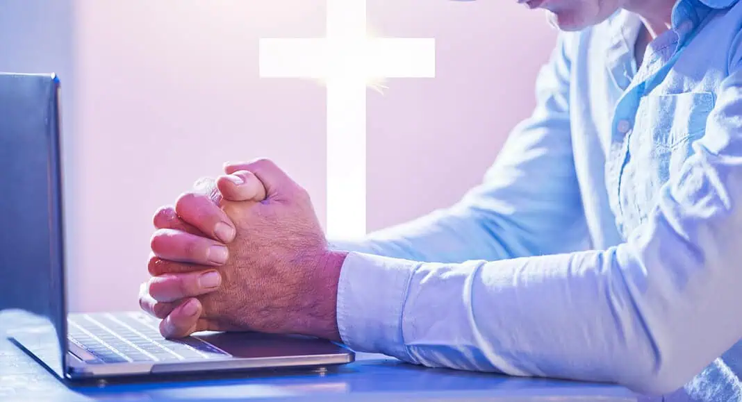 Un uomo che indossa una cuffia VR prega davanti a un computer portatile, interagendo con un chatbot AI, mentre sullo sfondo brilla una croce bianca luminosa.