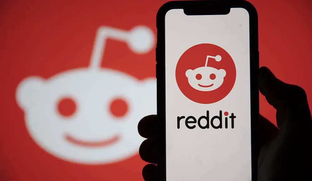 Una mano regge un telefono che mostra il logo di Reddit, con un logo di Reddit più grande sullo sfondo, evidenziando la popolare app Reddit.