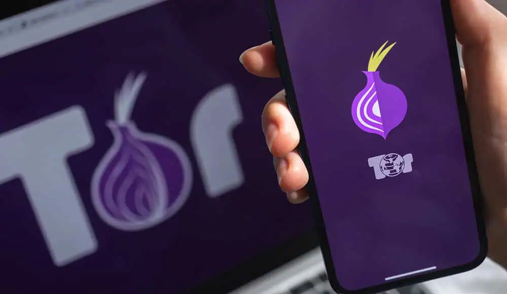 Una mano regge un telefono che mostra il logo di Tor Browser, mentre lo stesso logo di Tor Browser appare sullo schermo di un computer portatile sullo sfondo.