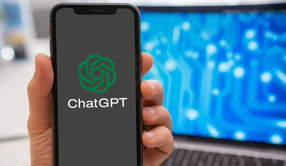 Una mano regge uno smartphone che mostra il logo ChatGPT, con un computer portatile e un circuito sullo sfondo, evidenziando l'integrazione di ChatGPT nella tecnologia moderna.