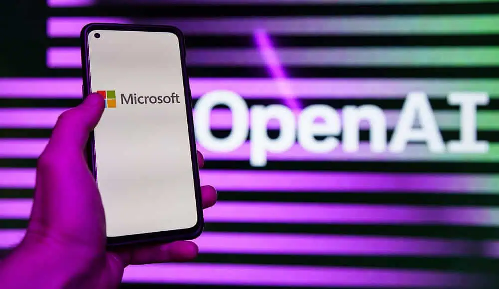 Una mano regge un telefono con il logo Microsoft; il nome di OpenAI si illumina di viola sullo sfondo.