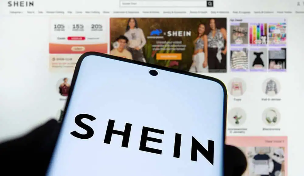 Uno smartphone mostra il logo Shein davanti alla homepage del sito di shopping online Shein, evidenziando la presenza digitale del marchio.