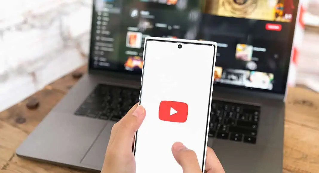 Una persona tiene un telefono con l'applicazione YouTube aperta davanti a un computer portatile, entrambi gli schermi visualizzano i contenuti di YouTube.