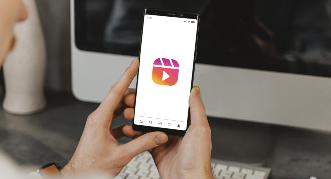 Persona che tiene in mano uno smartphone con il logo di Instagram Reels, con un computer e una tastiera sullo sfondo, per evidenziare le possibilità creative di Instagram.