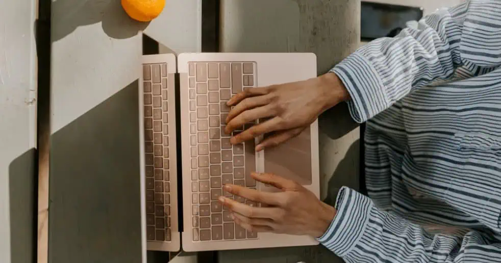 Persona con camicia a righe che scrive su un computer portatile con Windows 11 a un tavolo di legno, con un'arancia a lato.