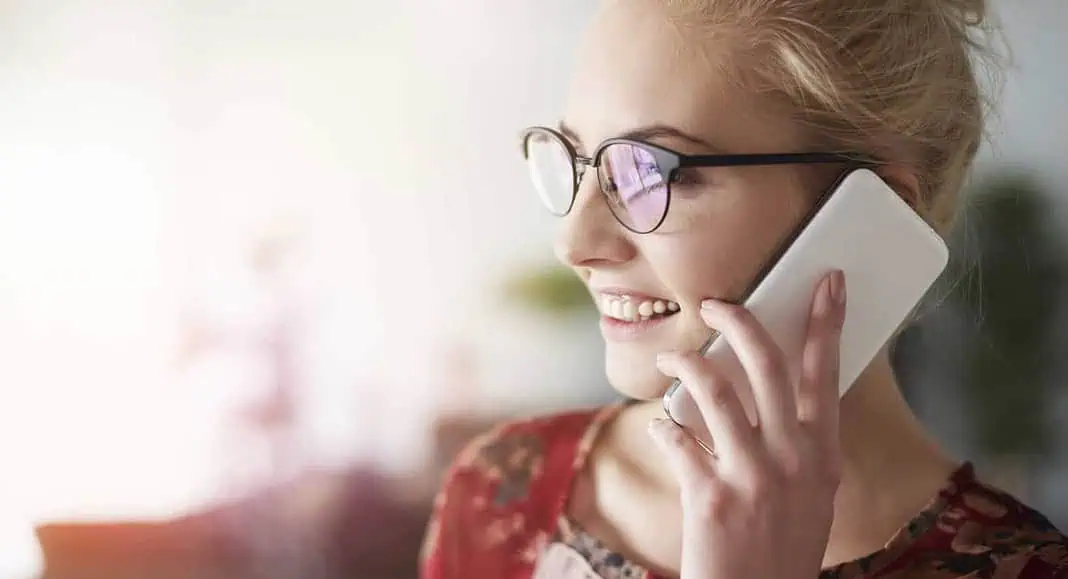 Una donna sorridente con gli occhiali parla con uno smartphone, discutendo di tariffe telefoniche, con la luce del sole sullo sfondo.