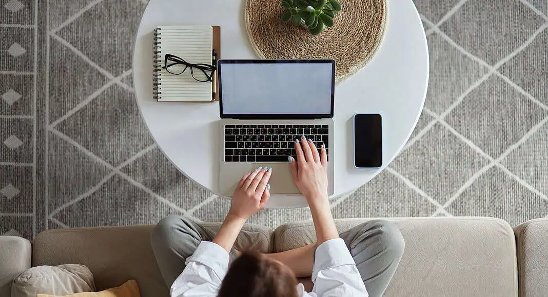 Persona che utilizza un computer portatile su un tavolo rotondo con un notebook, un telefono, la homepage di Google sullo schermo, occhiali e una pianta su un tappeto a motivi.