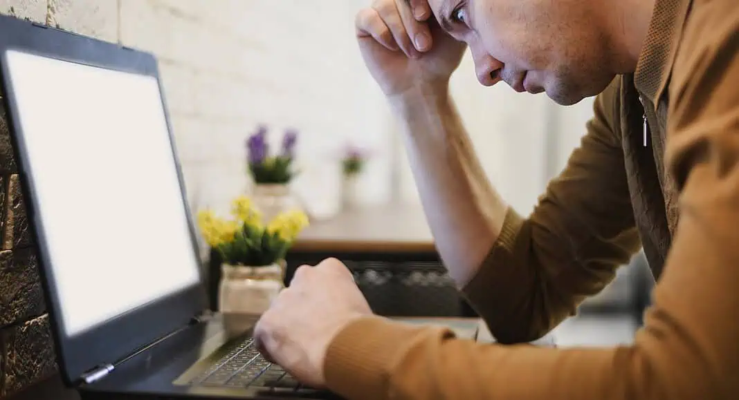 Un uomo sembra stressato mentre fissa lo schermo vuoto di un computer portatile Windows 11 in una stanza accogliente con piccole piante in vaso.