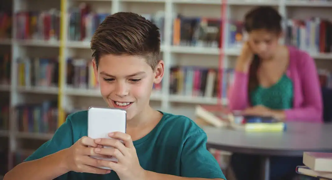 Ragazzo sorridente che utilizza uno smartphone in una biblioteca con libri, uno studente che legge sullo sfondo e un poster luminoso di Comet visibile sulla parete.