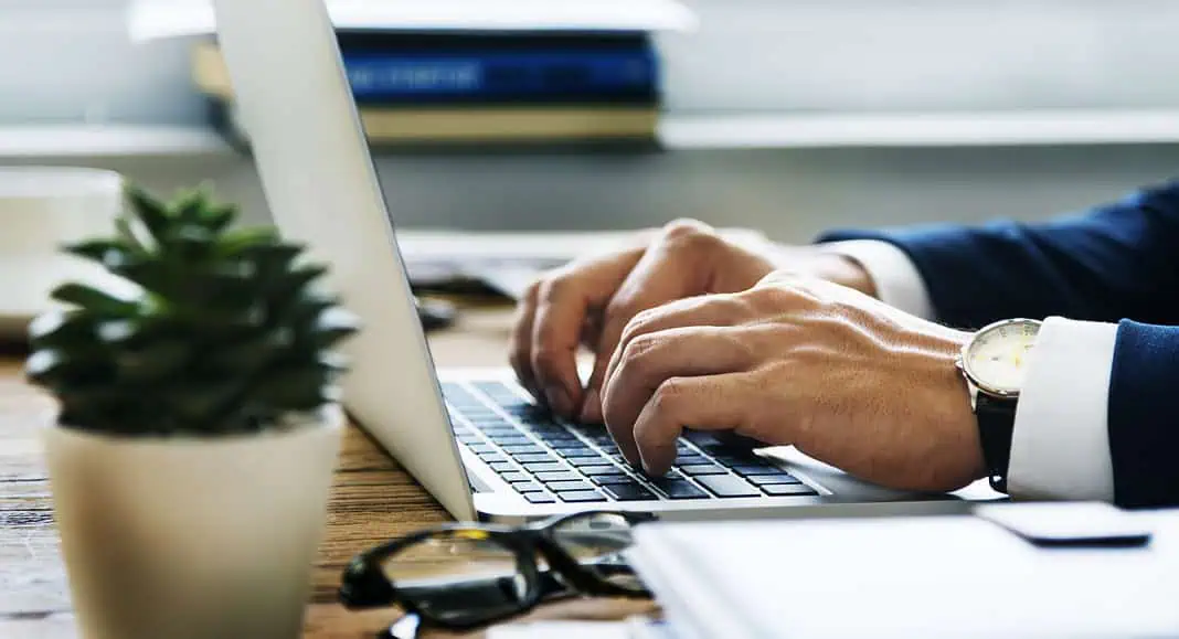 Primo piano delle mani di un giocatore A che scrive su un computer portatile a una scrivania, con una pianta e dei libri sullo sfondo.