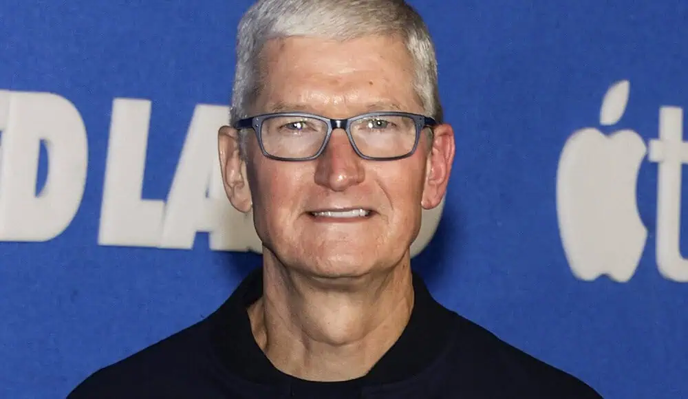 Un uomo con capelli corti e occhiali sorride davanti a uno sfondo blu con l'iconico logo Apple.