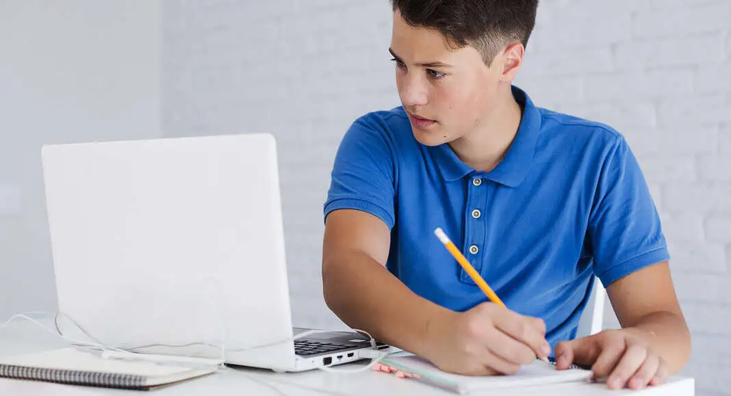 Ragazzo in camicia blu che scrive su un taccuino guardando un computer portatile su una scrivania bianca, utilizzando ChatGPT per un aiuto allo studio.