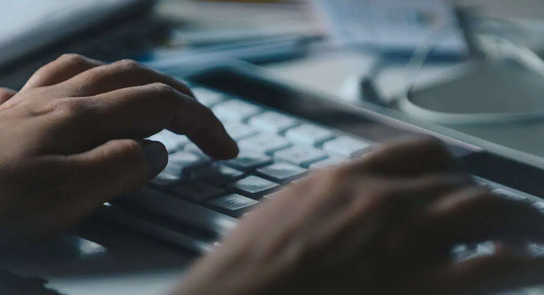 Mani che digitano su una tastiera bianca, con documenti e cavi sfocati sullo sfondo, che suggeriscono un lavoro sulle soluzioni cloud AWS.