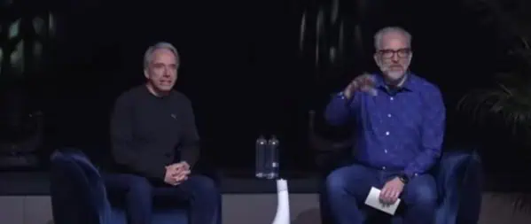 Due uomini siedono sul palco su sedie, uno dei quali regge dei fogli, con uno sfondo scuro e due bottiglie su un tavolino, che ricordano le interviste a pionieri della tecnologia come Linus Torvalds.