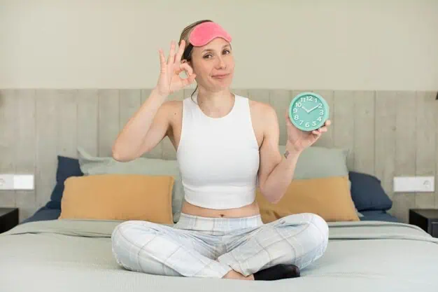 Donna in pigiama seduta sul letto, con un orologio in mano e un segno di OK, con una mascherina rosa in testa.